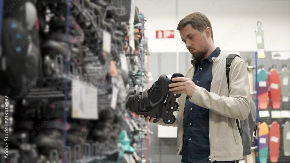 Young guy with a beard wants to buy brand new roller skates for skating in summer. Man wants to get fit doing cardio on roller skates. Sport shop. Young guy with a beard wants to buy brand new roller skates for skating in summer. Man wants to get fit doing cardio on roller skates. Sport shop.