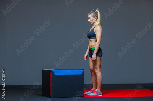 Beautiful female fitness athlete performs box jumps in a dark gym wearing black sports top and short tights with face hidden.