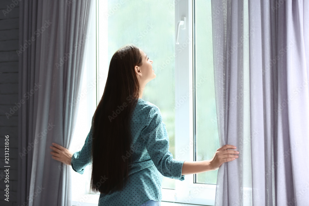 Young woman opening curtains in room near big beautiful window Young woman opening curtains in room near big beautiful window