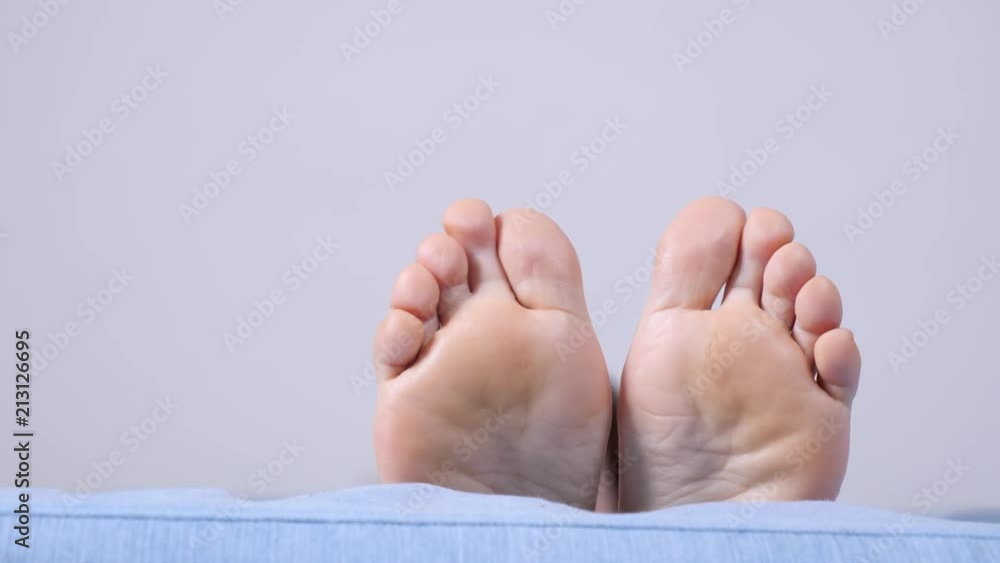 Female resting on sofa showing bare foot. Human feet moving on bed in slow motion, white grey wall background copy space text area. Female resting on sofa showing bare foot. Human feet moving on bed in slow motion, white grey wall background copy space text area.