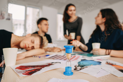 time management, deadline, sleep deprivation, daily routine. young worker sleeping at work during business meeting, hourglass on the fore plane