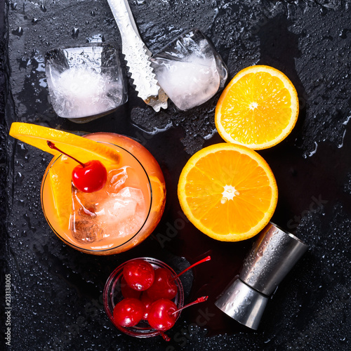 Tequila Sunrise cocktail, orange, ice cubes, maraschino cherries, ice tongs and jigger on a wet black slate tray. Cocktail Set. Top view. photo