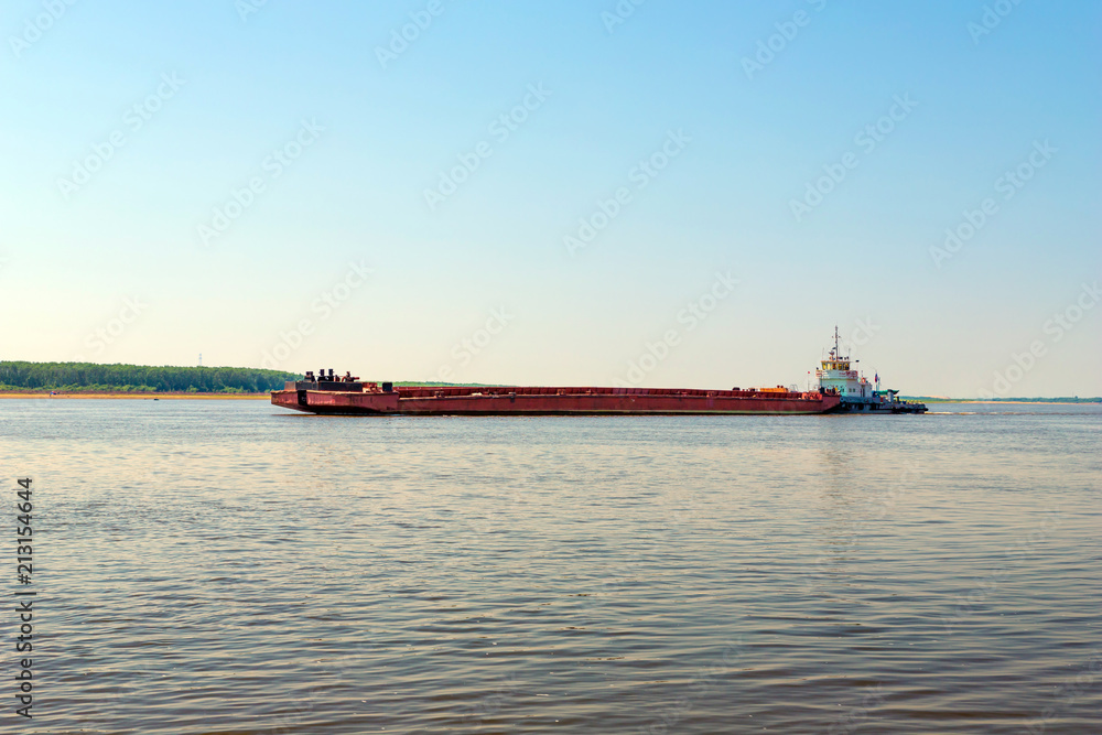 The tugboat pulls an empty barge in front of him. A river cargo ship sails along a wide river in a bright sunny day. Small hills covered by forest at the background.
