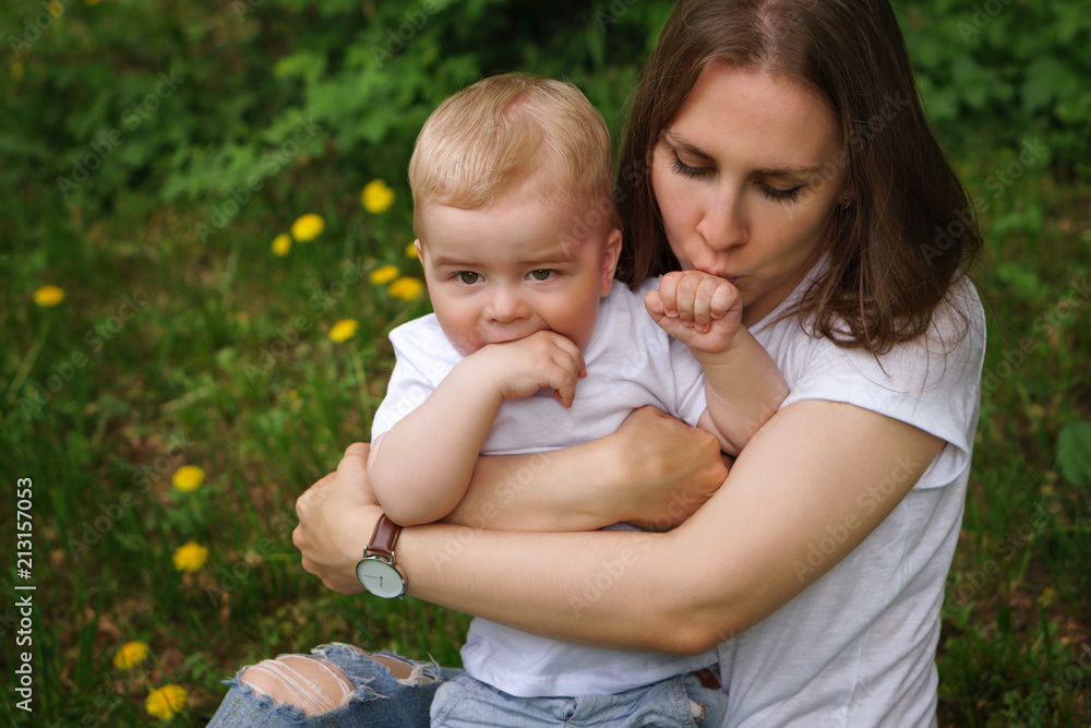 Young pregnant girl sits on lawn in city park. Mother hugs and kisses her son's hand. Little boy bites his hand. Happy motherhood Young pregnant girl sits on lawn in city park. Mother hugs and kisses her son's hand. Little boy bites his hand. Happy motherhood