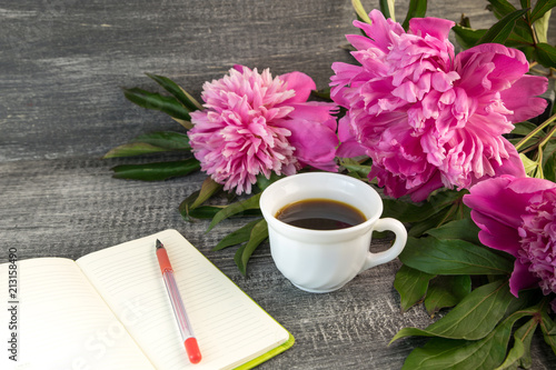 White cup of coffee with bouquet of large bright pink peonies with notebook and red pen on a grey wood table.