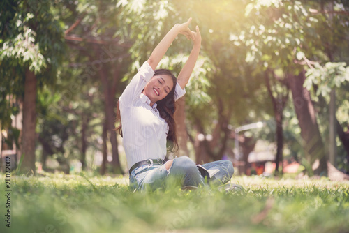 Beautiful Asian young woman sitting and relaxing on the field in green grass, and enjoy nature in park. Healthy Smiling Girl breathing fresh air,