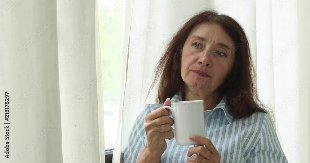 Middle-aged woman with brown hair wearing striped shirt and holding cup of coffee looking away in contemplation Middle-aged woman with brown hair wearing striped shirt and holding cup of coffee looking away in contemplation
