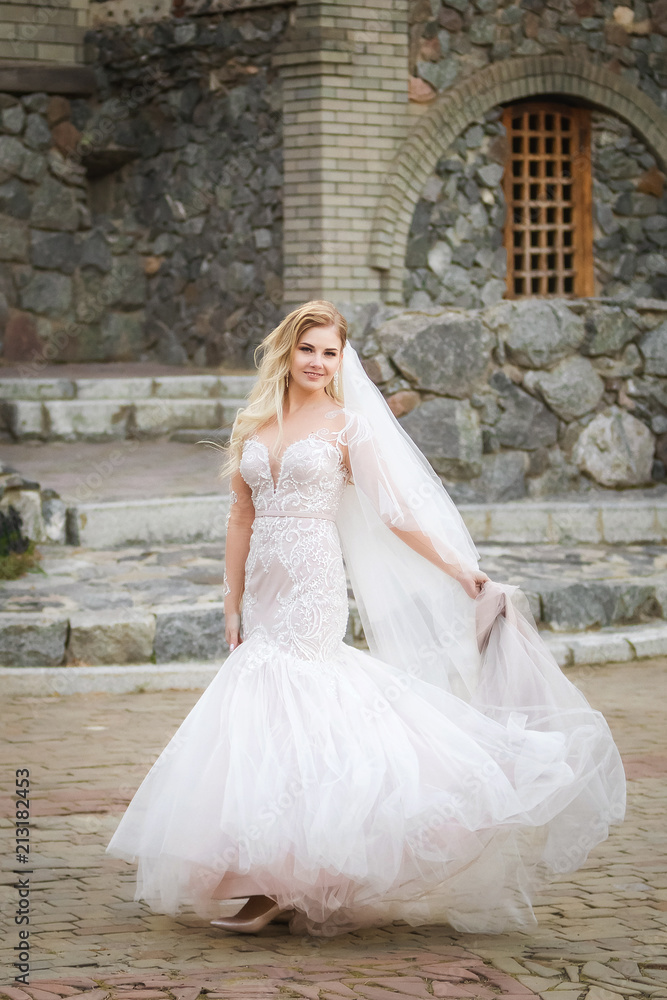 Beautiful sexy smiling bride in elegant wedding dress keeping long skirt near the old historical building