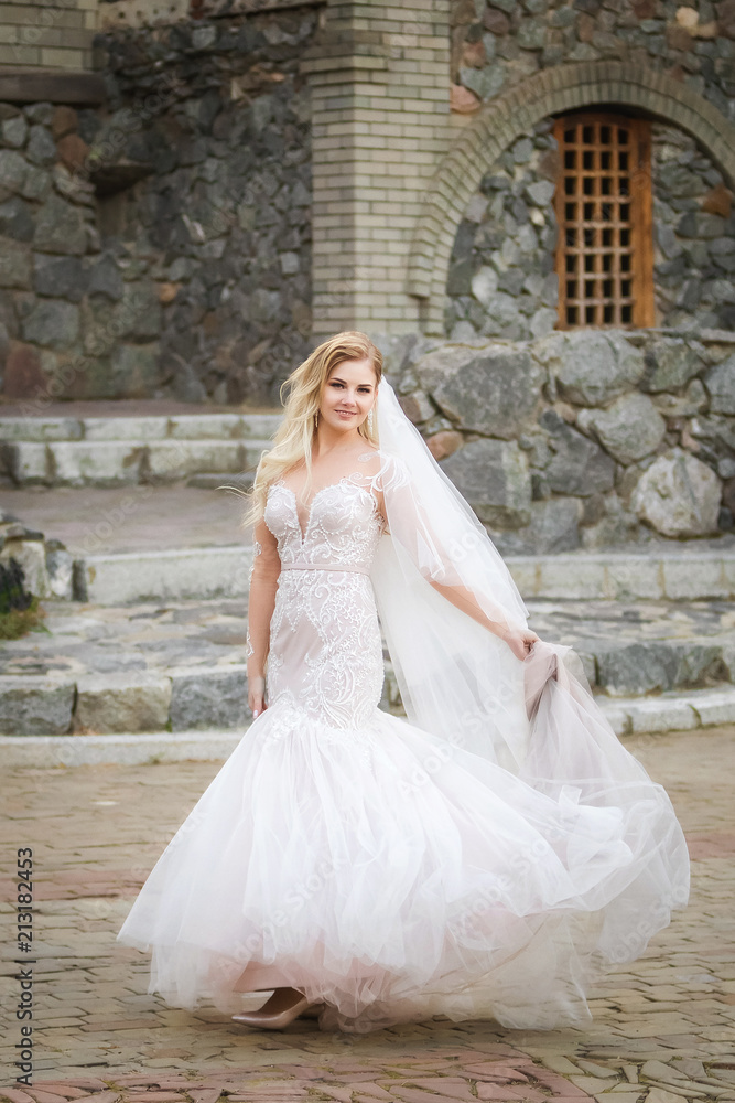 Beautiful sexy smiling bride in elegant wedding dress keeping long skirt near the old historical building Beautiful sexy smiling bride in elegant wedding dress keeping long skirt near the old historical building