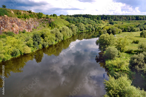 two banks of the river: rocky and plain, covered with green grass and dense trees, in sunny weather under a blue cloudy sky. resting place, fishing and picnic © adamchuk_leo