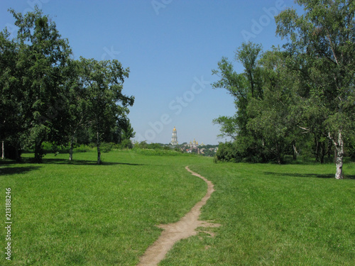 The path leading through the park area with a green lawn and tall deciduous trees against the backdrop of a beautiful temple in the distance