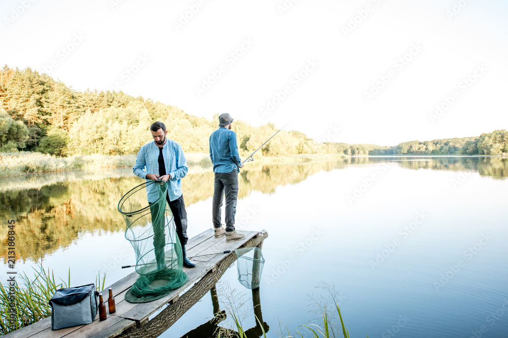 Two male friends preparing for fishing standing with fishing net and rod on the wooden pier during the morning light on the lake