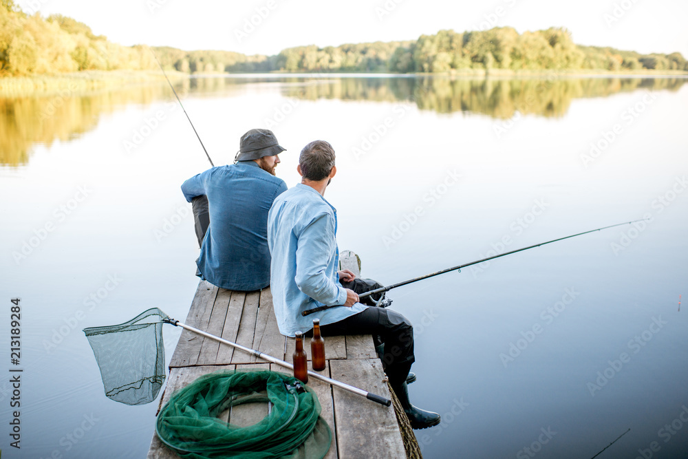 Two male friends dressed in blue shirts fishing together with net and rod  sitting on the wooden pier during the morning light on the lake Stock Photo