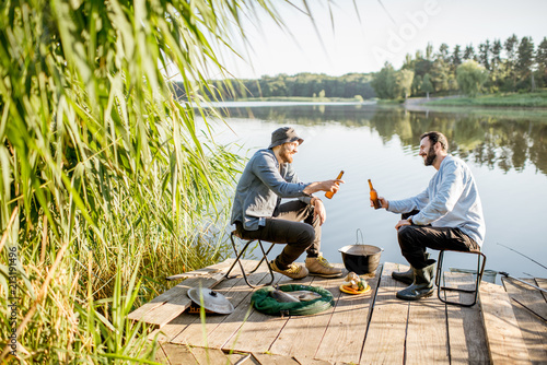 Two male friends relaxing with beer sitting together during the fishing process on the pier near the lake