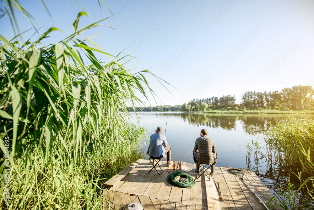 Landscape view on the beautiful lake and green reeds with two men fishing on the wooden pier during the morning light Landscape view on the beautiful lake and green reeds with two men fishing on the wooden pier during the morning light