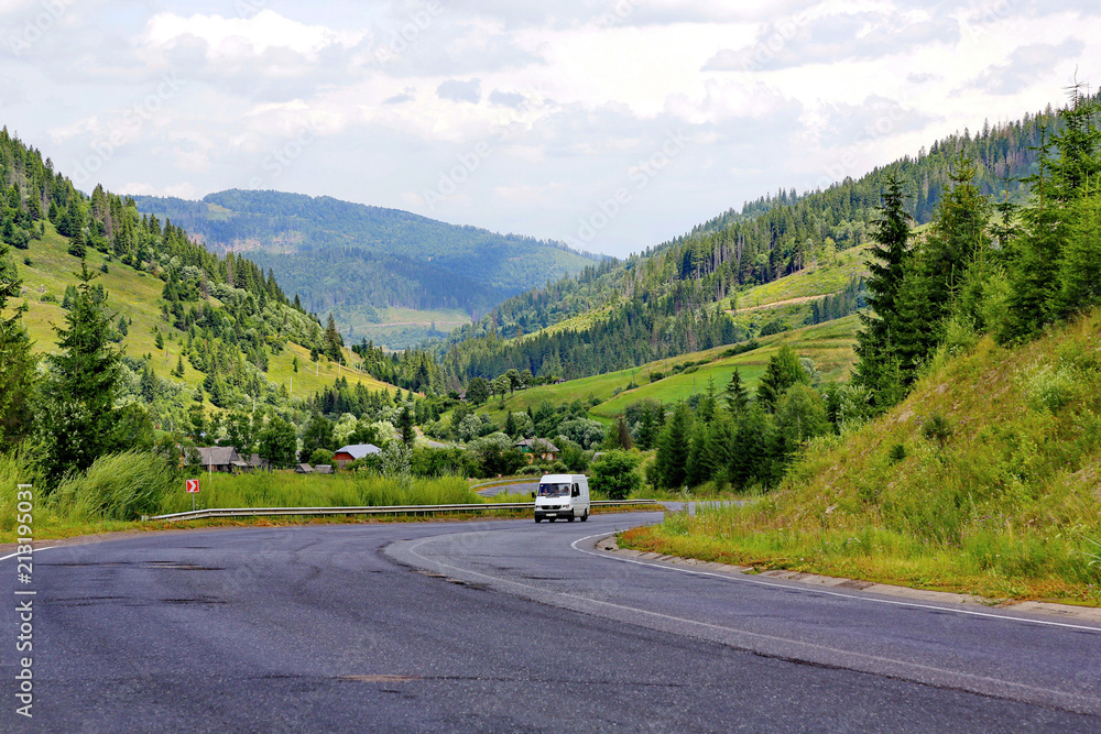 Fototapeta premium The car rides between the high green mountain slopes along the road winding between them. With the roofs of rural houses seen among the trees standing on the roadside.
