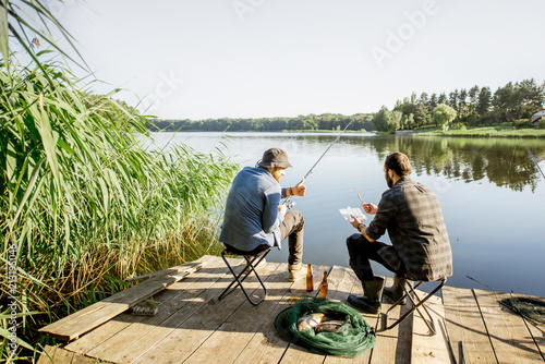 Landscape view on the beautiful lake and green reeds with two men fishing on the wooden pier during the morning light