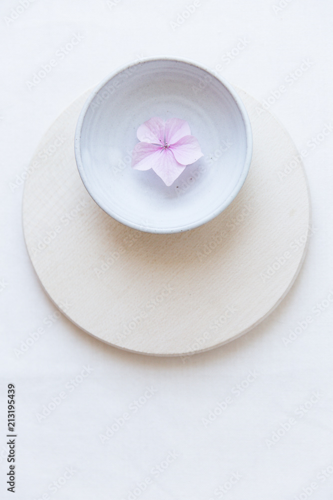 A single pink hydrangea flower in a small ceramic bowl on a round wooden plate on a white surface seen from above. A single pink hydrangea flower in a small ceramic bowl on a round wooden plate on a white surface seen from above.