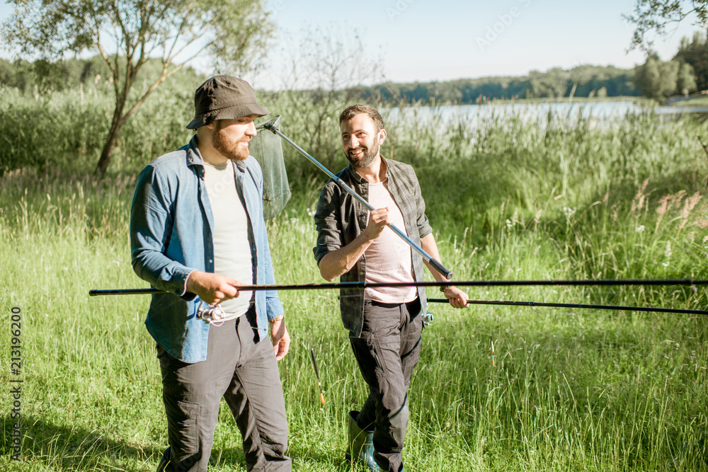 Two fishermen walking with fishing rod and net on the green lawn near the lake in the morning Two fishermen walking with fishing rod and net on the green lawn near the lake in the morning