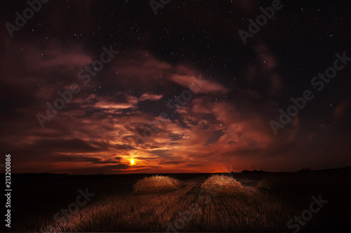 Night photo. Straws of straw on the cleaned field and starry sky with clouds and moon. 