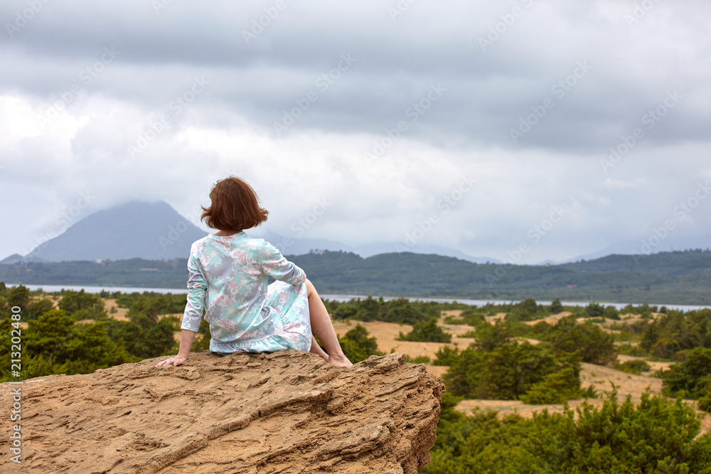 Young woman sits on a rock against the blue sea, yellow sand and sky with clouds and admires nature, enjoying solitude