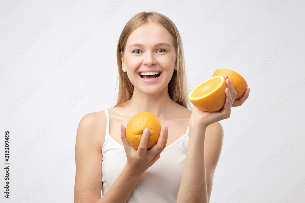 Beautiful young blonde woman in white shirt holding piece of orange near her head smiling while standing against white background. Concept of eating fruits and healthy diet Beautiful young blonde woman in white shirt holding piece of orange near her head smiling while standing against white background. Concept of eating fruits and healthy diet