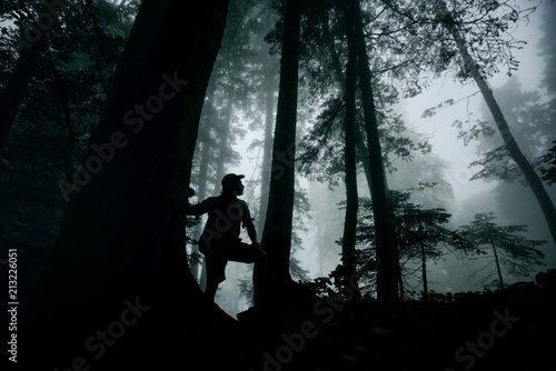 Silhouette of a traveler in a hat and with a backpack in a mysterious forest in the background of fog and trees. Man, freedom, travel.