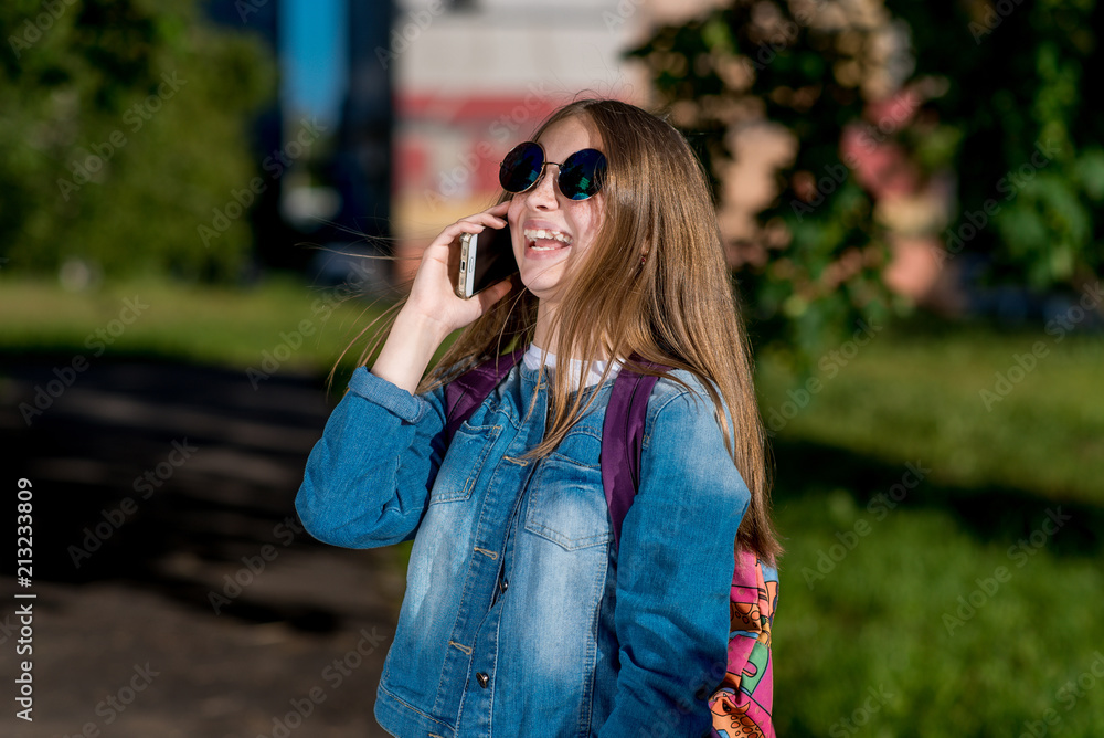 Girl schoolgirl talking smartphone. In summer, in denim clothing, a backpack in sunglasses is behind her back. He laughs brightly and emotionally. Smiles and enjoys the conversation. Girl schoolgirl talking smartphone. In summer, in denim clothing, a backpack in sunglasses is behind her back. He laughs brightly and emotionally. Smiles and enjoys the conversation.