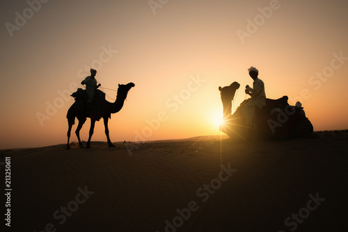 Rajasthan travel background - Three indian cameleers  camel drivers  with camels silhouettes in dunes of Thar desert on sunset. Jaisalmer  Rajasthan  India