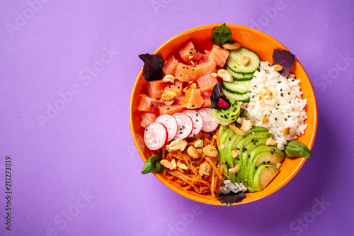 Hawaiian salad Poke with salmon fish, avocado, Korean carrots, cucumbers, radish and rice, with black sesame. in an orange plate. On the purple trend background of 2018. copy space, top view