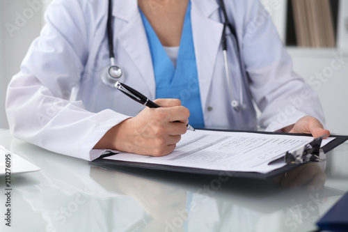 Doctor woman filling up medical form while sitting at the table, close-up of hands