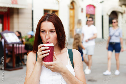 Young beautiful redhead woman drinking coffee from plastic cup walking down a street