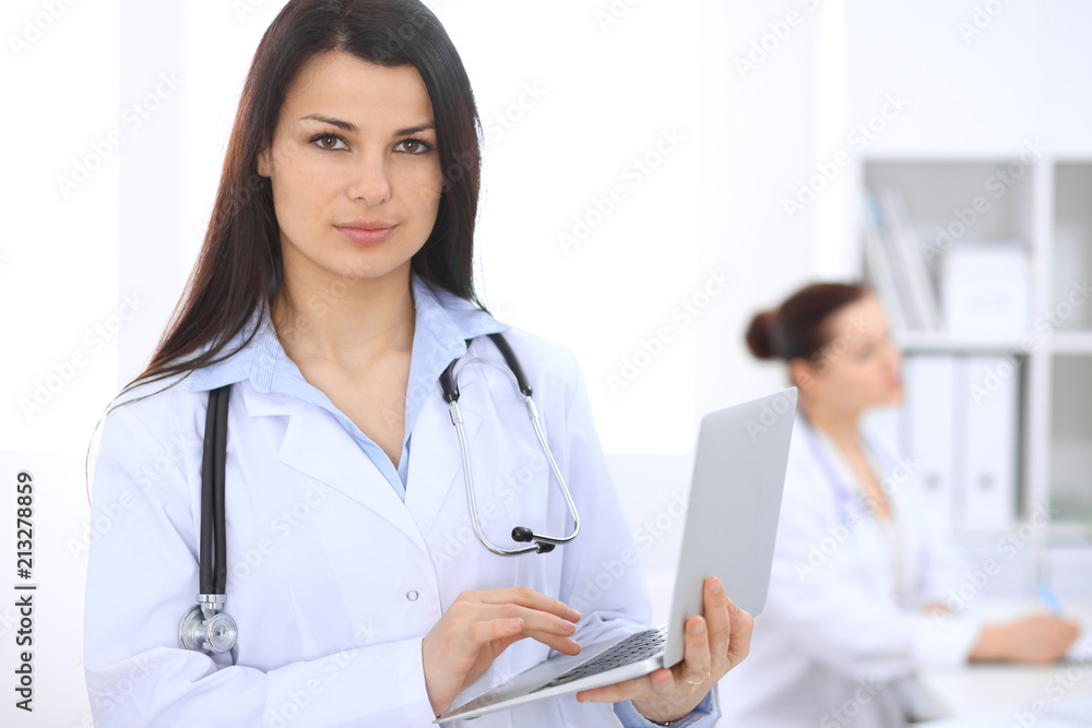 Brunette female doctor on the background of colleagues talking to each other in hospital. Medicine and health care concept Brunette female doctor on the background of colleagues talking to each other in hospital. Medicine and health care concept