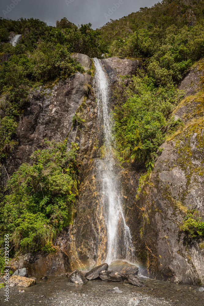 Obraz premium Waterfall creating a small rainbow and vegetation on a stormy day at Franz Josef Glacier, south island New Zealand