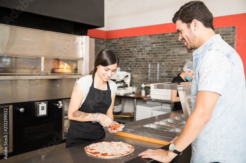 Smiling woman adding pepperoni slices to cheese pizza in kitchen counter