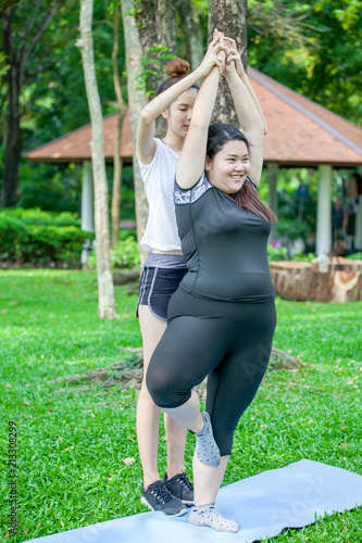 Foto de Asian Thin and overweight woman doing the tree yoga pose together  in park, fat and fitness girls yoga exercise training with assistance of  woman friend or personal trainer coach in