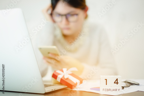 Business woman wearing glasses still working in Valentine day. Red gift box white ribbon using notebook laptop ,smartphone on office desk and wooden calendar 14 February. Hard work, holiday festival.