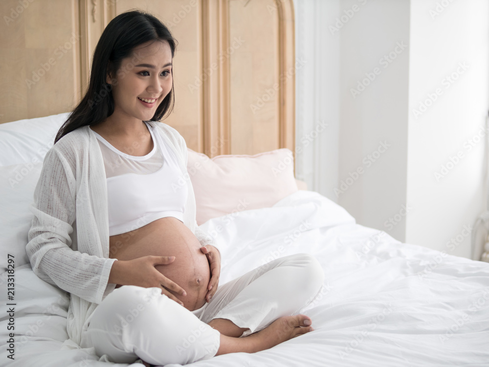 Happy pregnant asian woman sitting on bed and touching her belly in bedroom at home Happy pregnant asian woman sitting on bed and touching her belly in bedroom at home