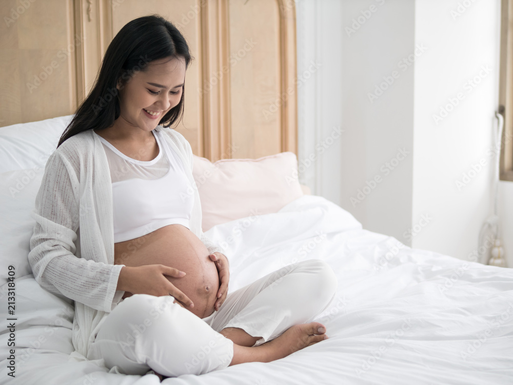 Happy pregnant asian woman sitting on bed and touching her belly in bedroom at home Happy pregnant asian woman sitting on bed and touching her belly in bedroom at home
