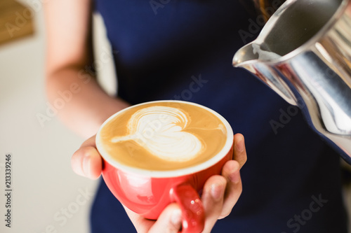 Barista pouring hot milk stream heart shape latte art on red cup coffee in vintage color tone.