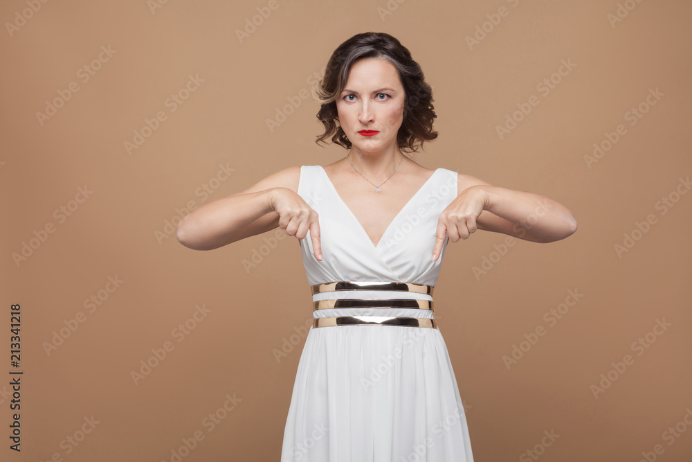 serious woman pointing finger down at copy space, look now! Emotional expressing woman in white dress, red lips and dark curly hairstyle. Studio shot, indoor, isolated on beige, light brown background serious woman pointing finger down at copy space, look now! Emotional expressing woman in white dress, red lips and dark curly hairstyle. Studio shot, indoor, isolated on beige, light brown background