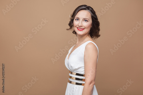 pretty middle aged toothy smiley woman looking at camera. Emotional expressing woman in white dress, red lips and dark curly hairstyle. Studio shot, indoor, isolated on beige or light brown background