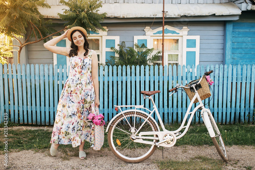 Smiling cute brunnete happy girl in a white-flowers dress and hat with pink peones. Lovely house is on the background photo