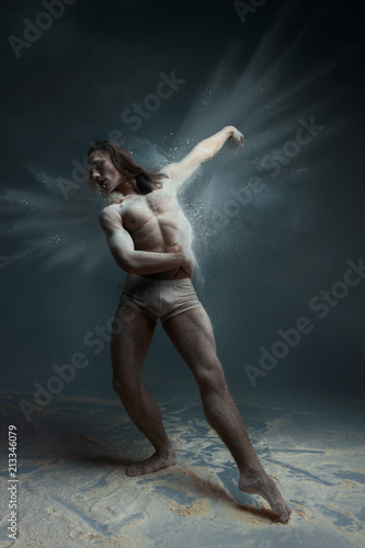 Dancing in flour concept. Long hair muscle fitness man male guy dancer in dust / fog. Guy wearing white shorts making dance element with foggy wings in flour cloud on isolated grey background