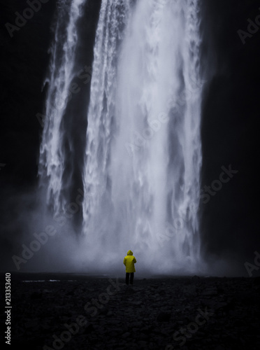 Famous Skogafoss Waterfall in Iceland with a small man in a yellow coat looking at the water falling off the hill in a dark look with black stones in the foreground and no people in front during night