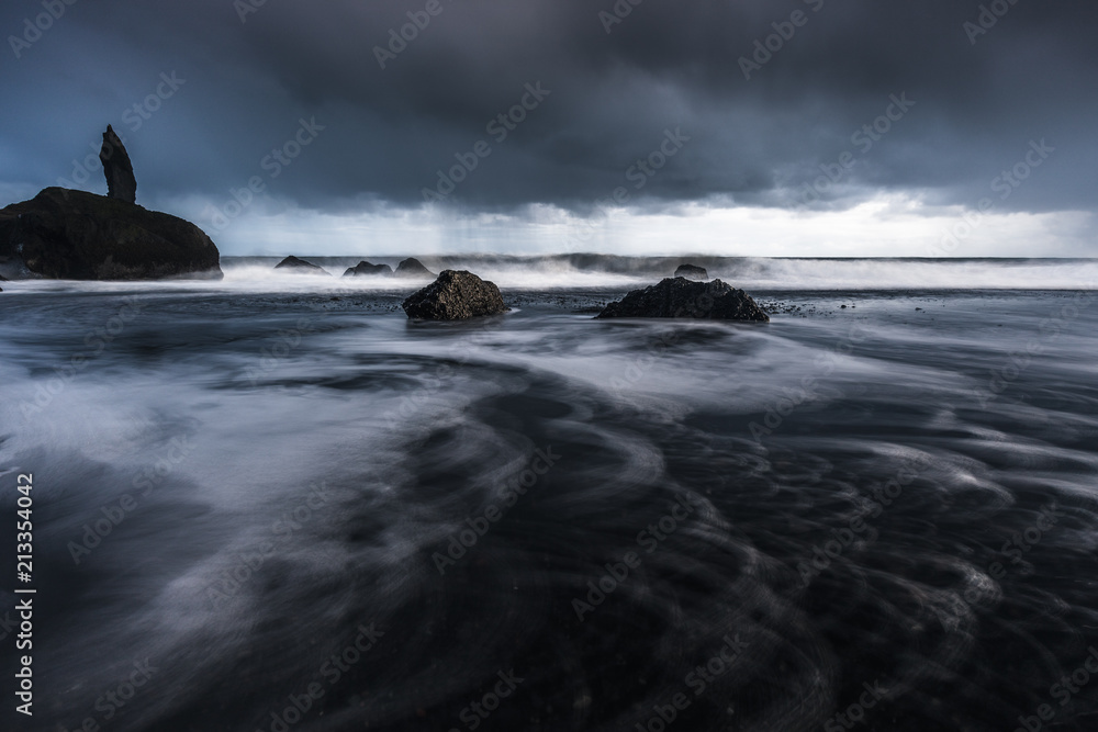 Rocks on a black sand beach in Iceland with reflection in the Sea and a dark sky in misty moody weather with dark colors and a rough sea in landscape format with a storm approaching