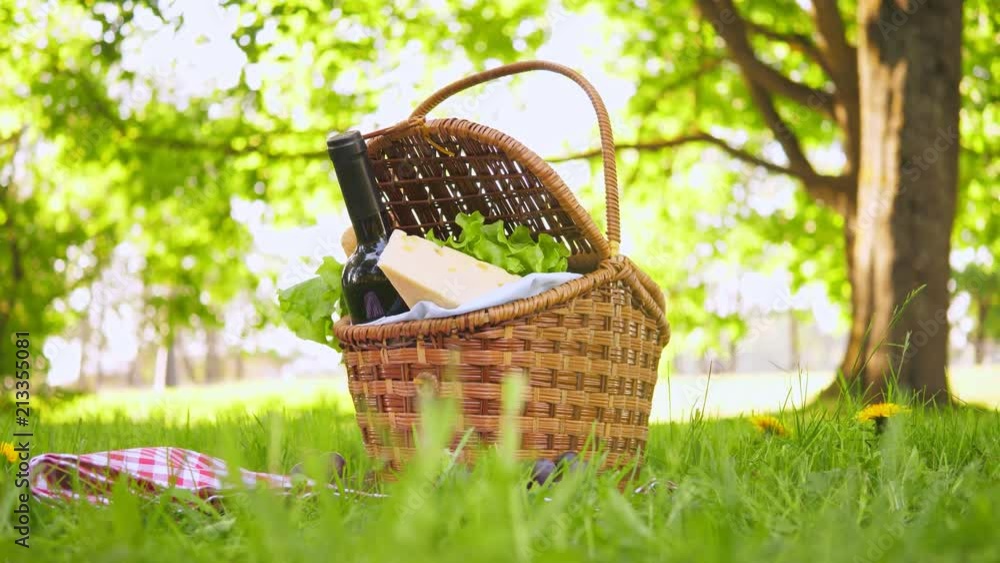 Wicker picnic basket with cheese and wine on red checkered table cloth on grass in park Wicker picnic basket with cheese and wine on red checkered table cloth on grass in park