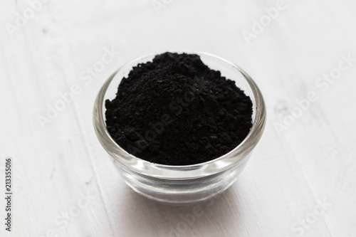 Activated charcoal powder in a glass bowl on white wooden background