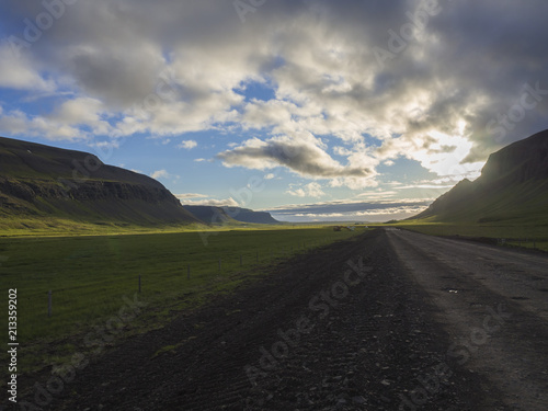 Dark dirty mountain road through valley in empty rural northern landscape with green grass and hills, water puddles and midnight sun, dramatic clouds, in Iceland west fjords, copy space