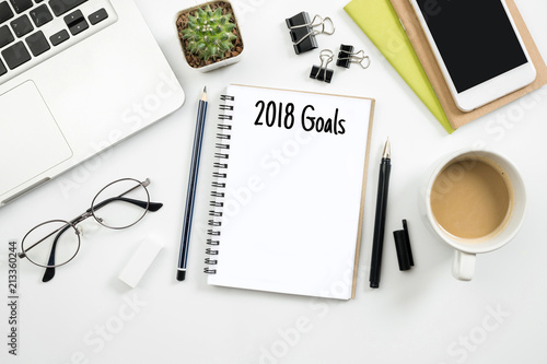 Notebook with 2018 goals text is on top of white office desk table. Top view, flat lay.